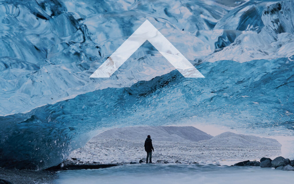 A person stands in the entrance of a huge, light-filled ice cave, looking out. Superimposed text: an upwards-pointing caret.