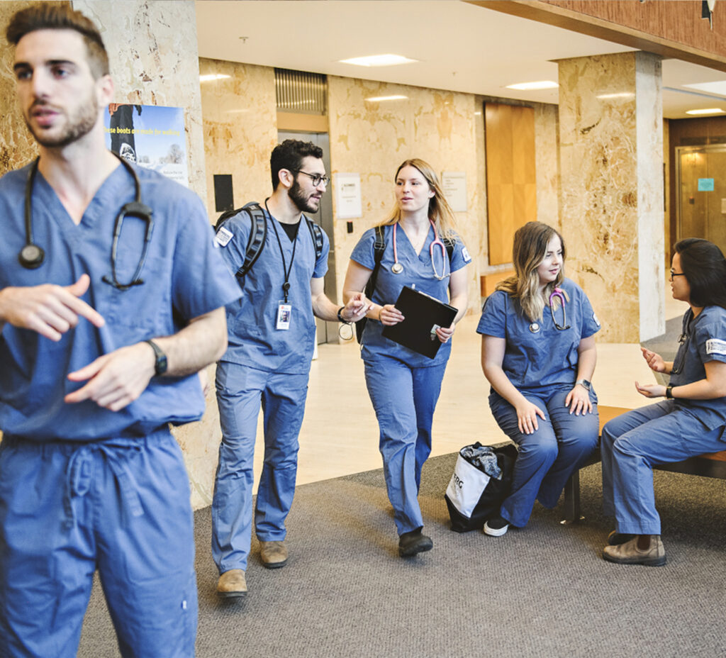 Five young people wearing nursing scrubs and stethoscopes chat as they exit through the lobby of a building.