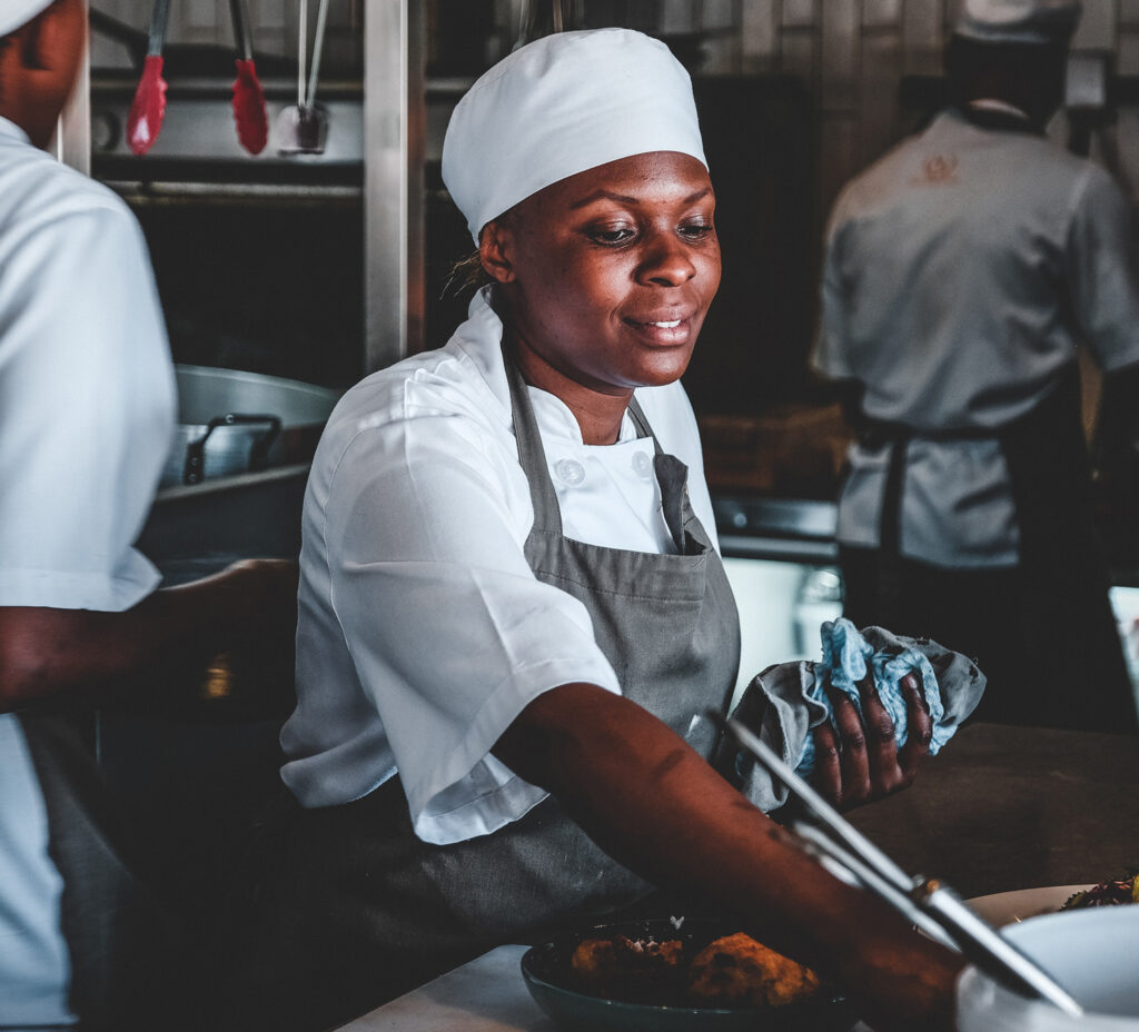 A Black woman wearing an apron and chef's cap works in a busy commercial kitchen.