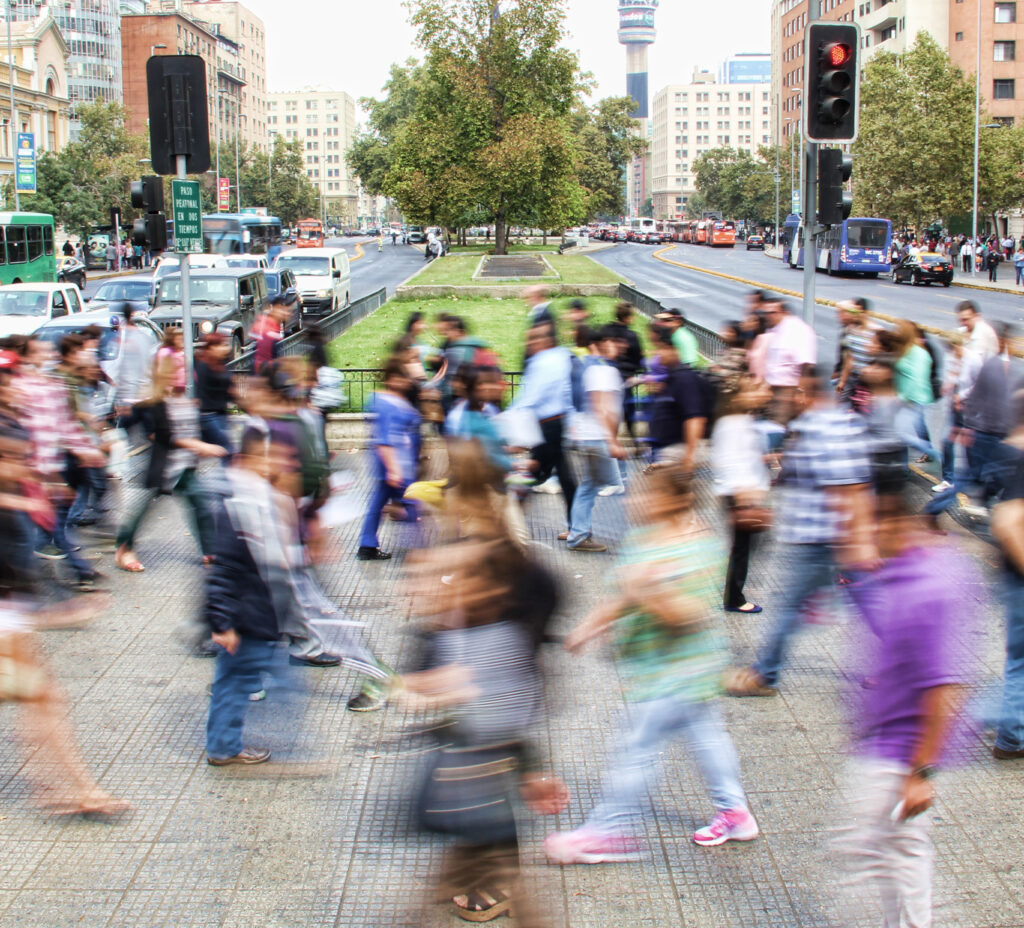 A crowd of pedestrians cross a busy street where a grassy median divides the opposing lanes of traffic.