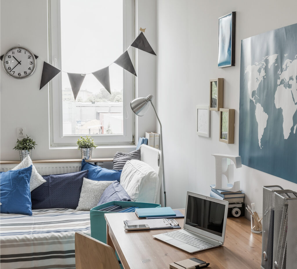 A room with a single bed, a desk with laptop, books and notebooks, a map on the wall, and pennants hung over the window.