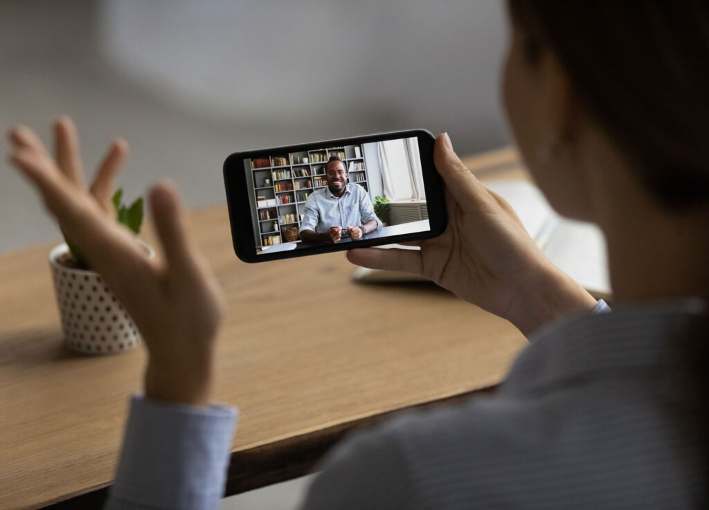 A woman holds a smartphone as she makes a video call with a man in a book-lined office.