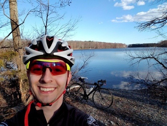 Adam Rosenfield, wearing bike helmet and sunglasses, stands on a lakeshore with his bike behind him.