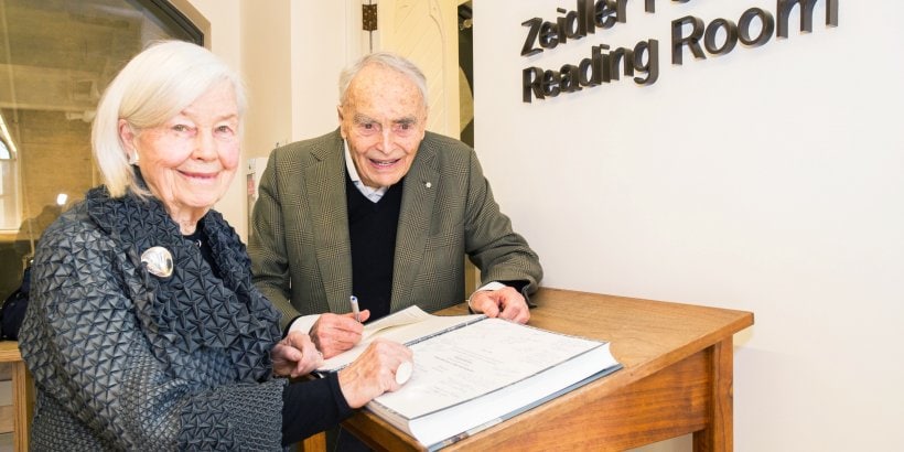 Jane and Eberhard Zeidler smile as they sign a guestbook. A sign on the wall says: Zeidler Family Reading Room.