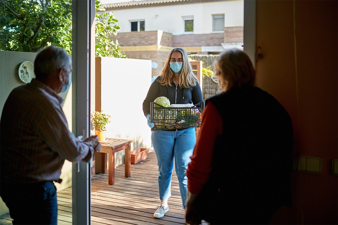 Two older people open their door to a young woman delivering groceries. Everyone wears a mask.