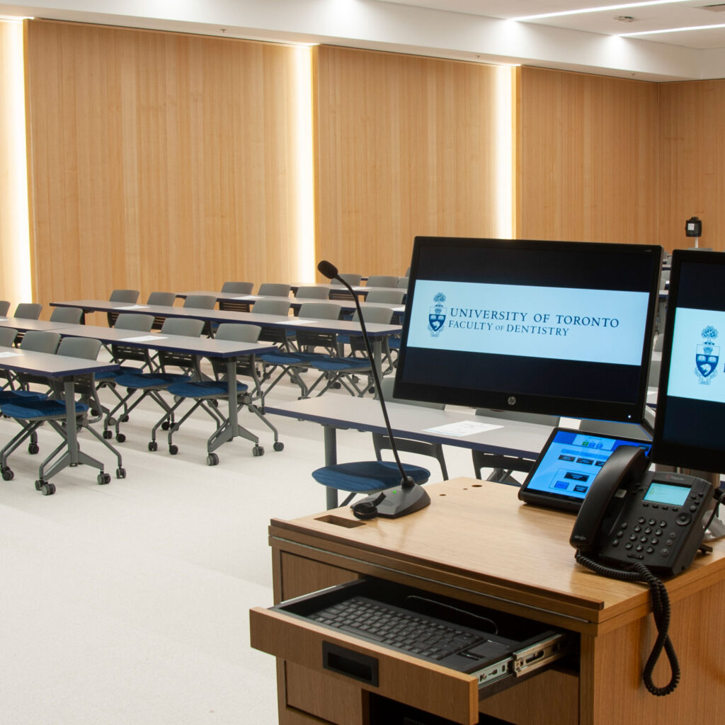 In the Faculty of Dentistry auditorium, tables and chairs face front. Lights shine from behind bright, wood panelled walls.