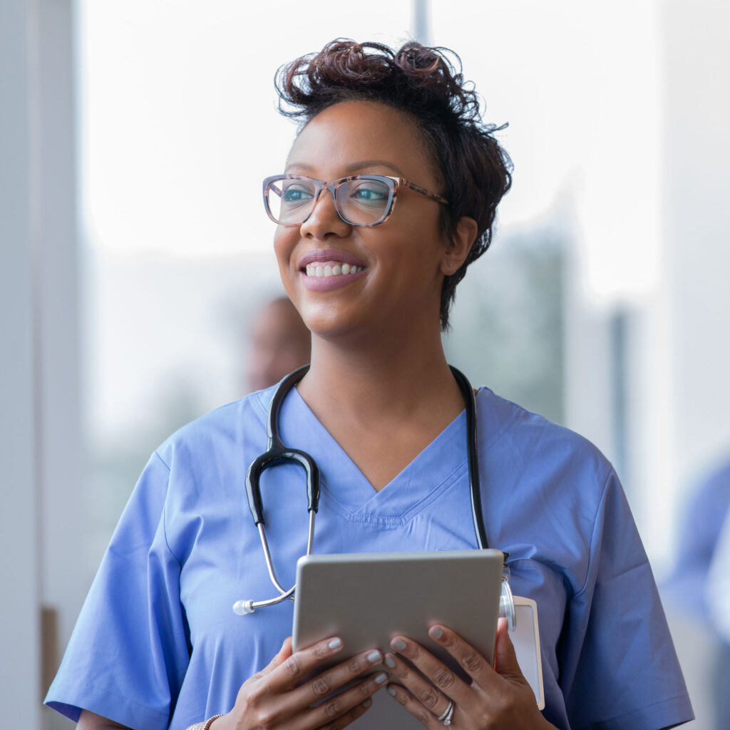 A smiling young Black woman, wearing nursing scrubs and holding a stethoscope and tablet.