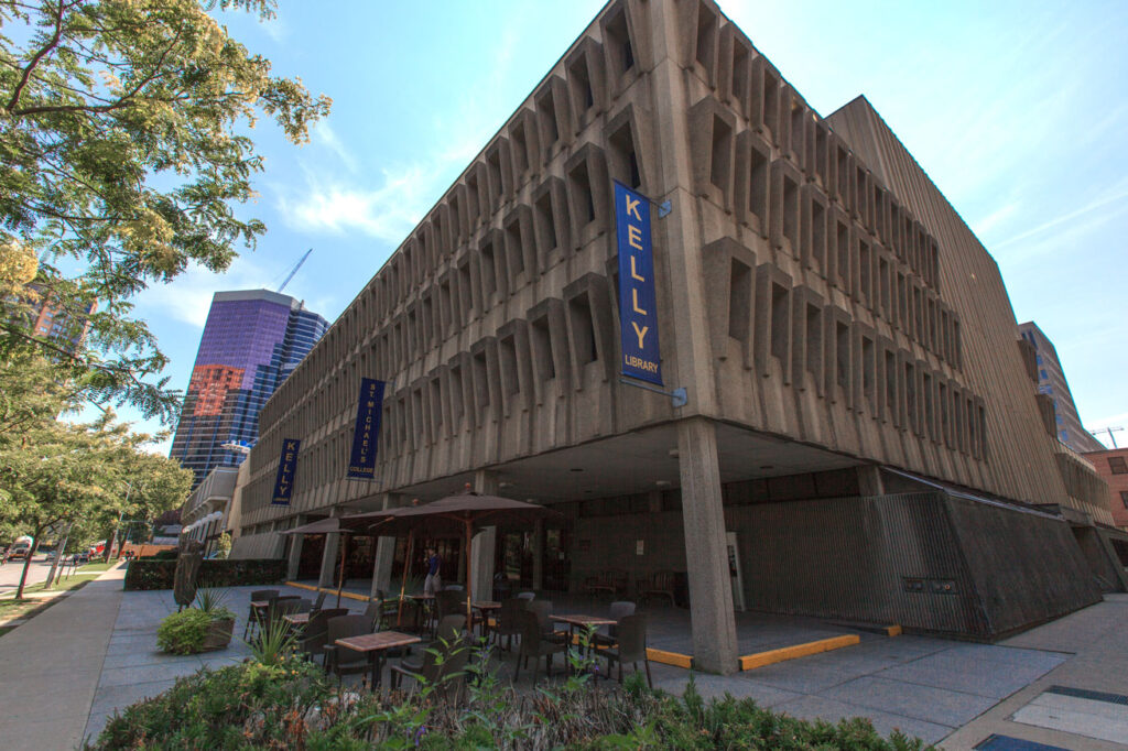 The Kelly Library has a concrete facade and an open portico. Blue banners hung from the building read: Kelly Library.