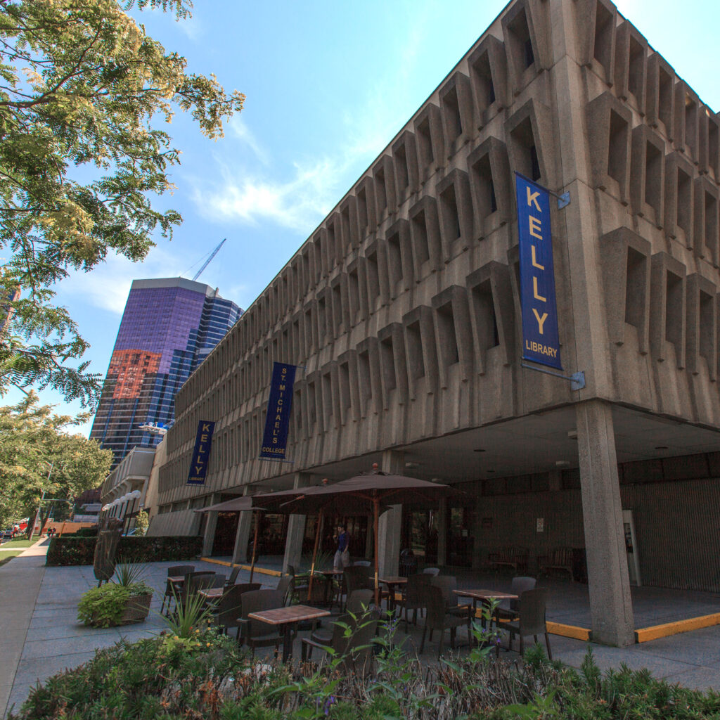 The Kelly Library has a concrete facade and an open portico. Blue banners hung from the building read: Kelly Library.