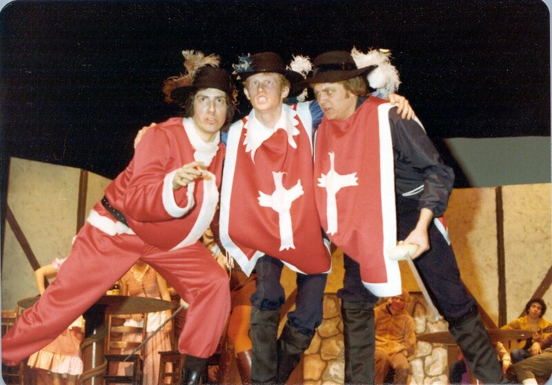 Three men on a stage huddle together. They wear 17th-century tabards and hats with feathers.