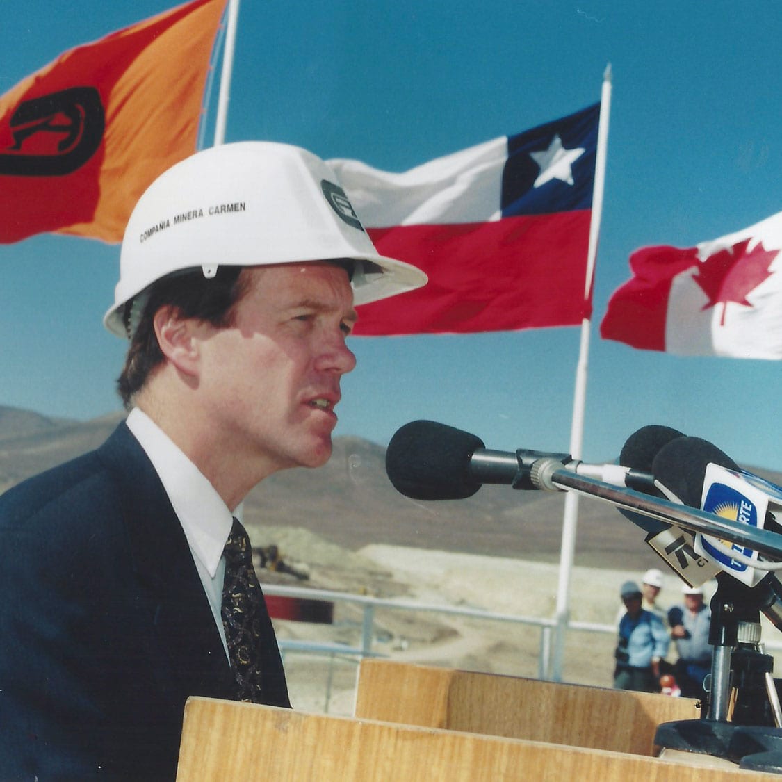 David Brace wears a hard hat and speaks into a microphone. Behind him are the flags of Chile and Canada.