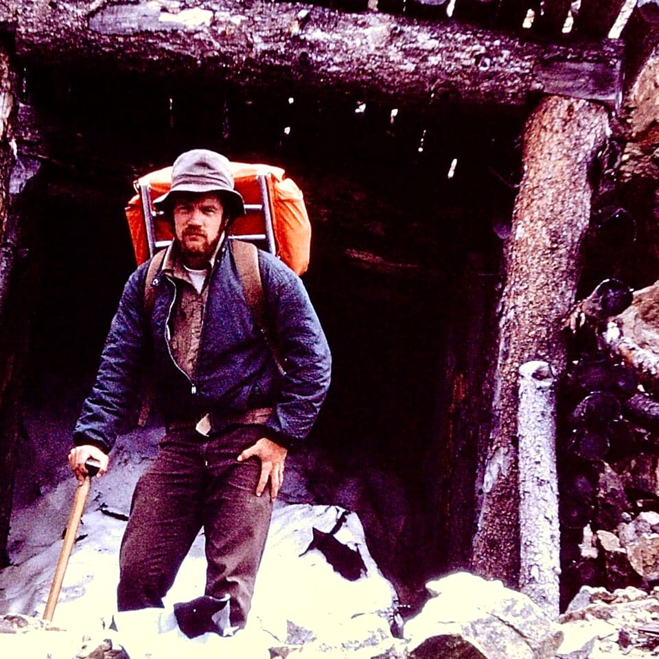 David Brace wears a huge backpack and stands in the snow outside a log shelter.