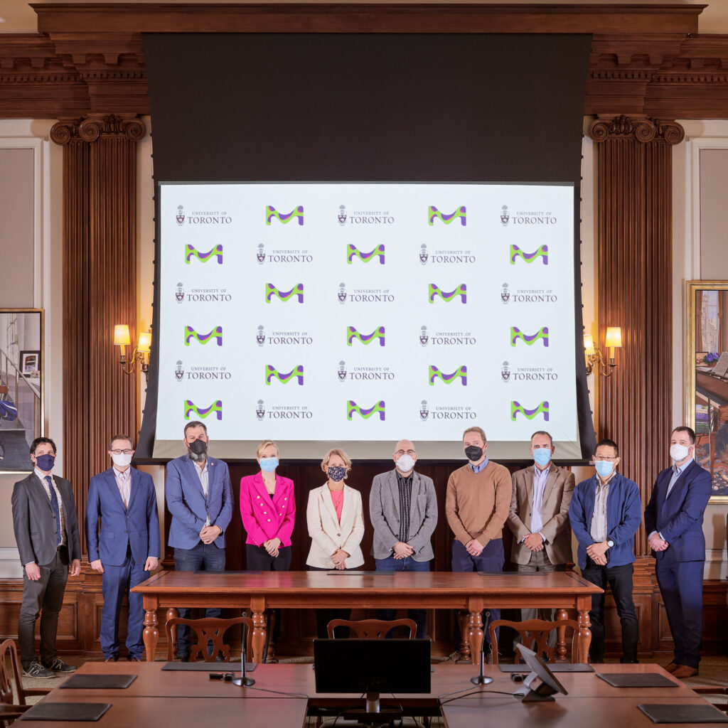 10 people pose for a photo in a boardroom, before a screen with the Merck KGaA and U of T logos.
