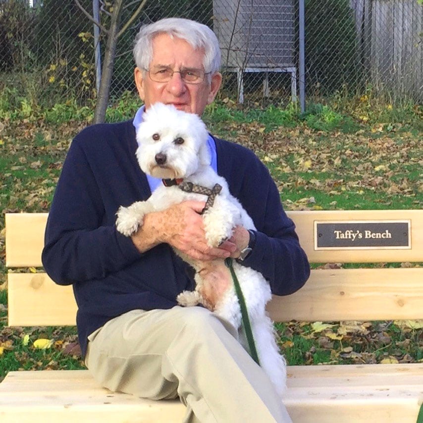 Edward Roberts sits on a bench, holding a small fluffy dog. Text on bench reads: Taffy's Bench.