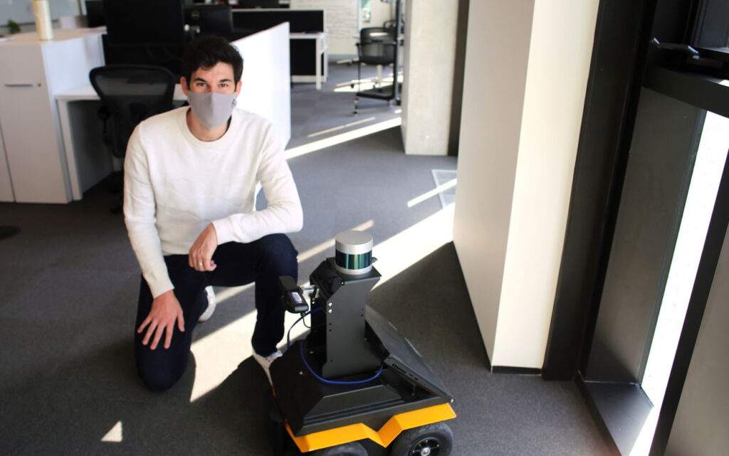 Hugues Thomas crouches beside a robot. It's made of a can-sized scanner and computer screen on top of a four-wheeled platform.