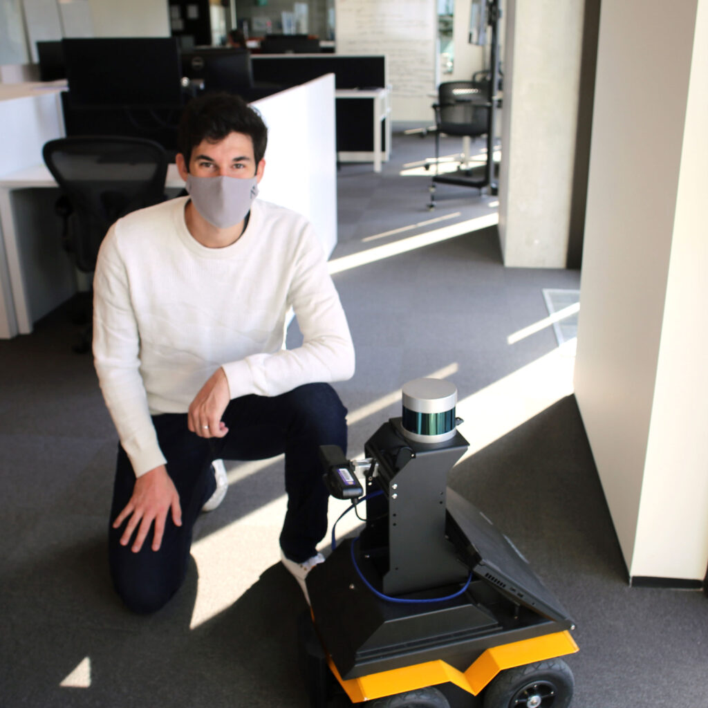 Hugues Thomas crouches beside a robot. It's made of a can-sized scanner and computer screen on top of a four-wheeled platform.