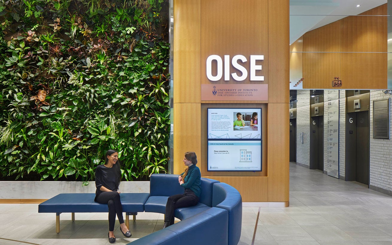 Two women relax on a bench in an atrium. Behind them, a lush vegetation wall and a sign that says, O.I.S.E.