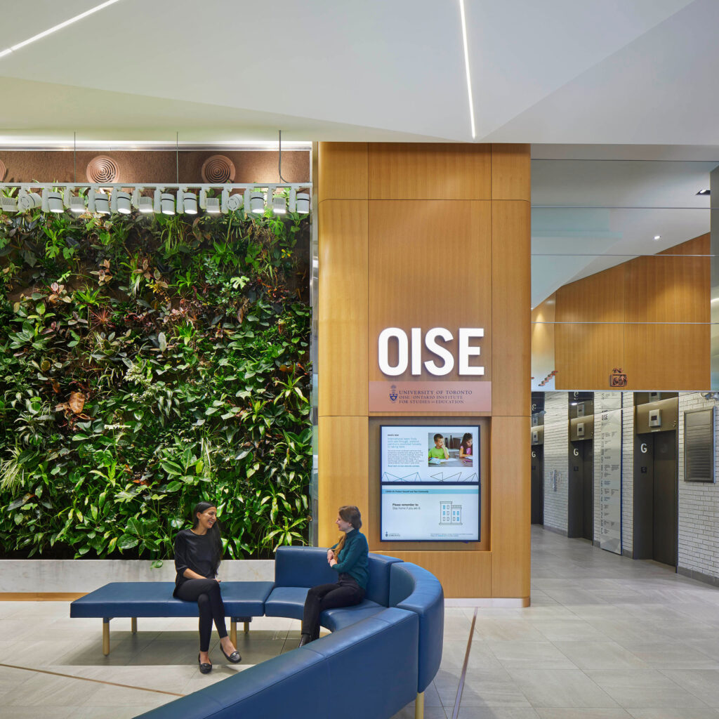 Two women relax on a bench in an atrium. Behind them, a lush vegetation wall and a sign that says, O.I.S.E.