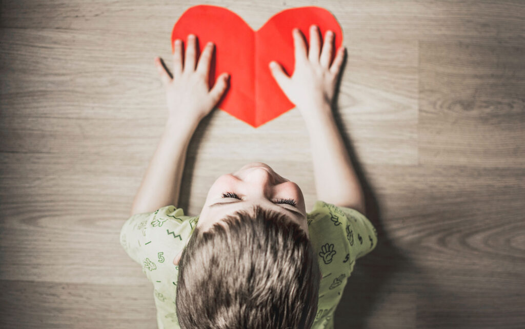 A young boy smiles as he lays out a big paper heart on a table.