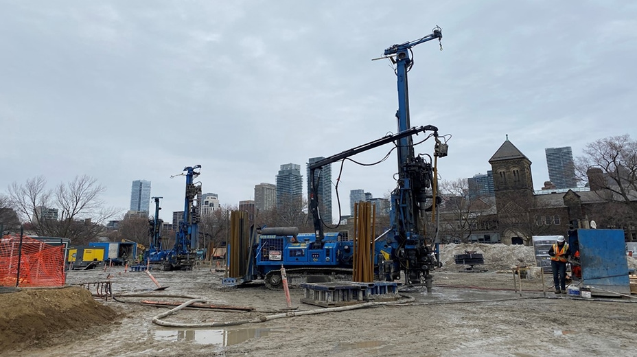 On a construction site, three cranes in a row are poised, holding large vertical shafts pointed at the ground.