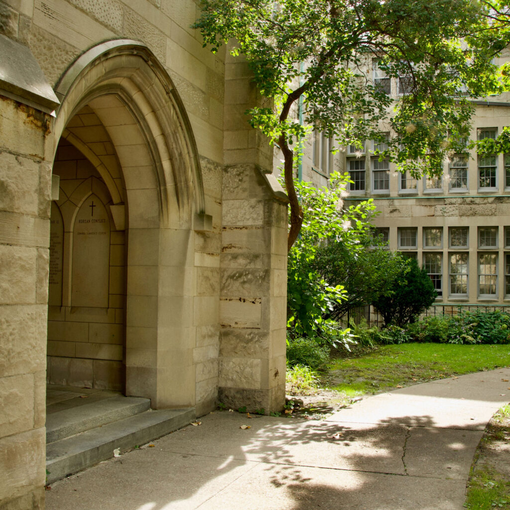 A green tree shades an old stone archway at St. Michael's College.