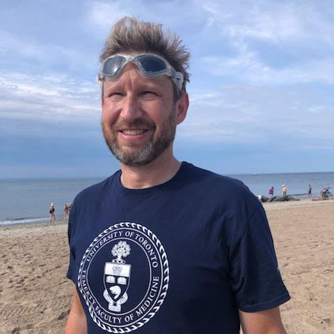 Sean Nuttall smiles, standing on a beach and wearing a Temerty Faculty of Medicine T-shirt.