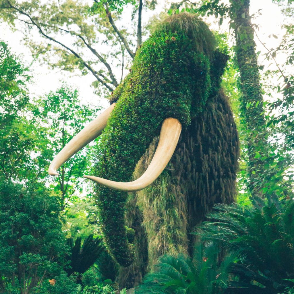 In a forest, shaggy plants grow over a framework in the shape of a woolly mammoth.
