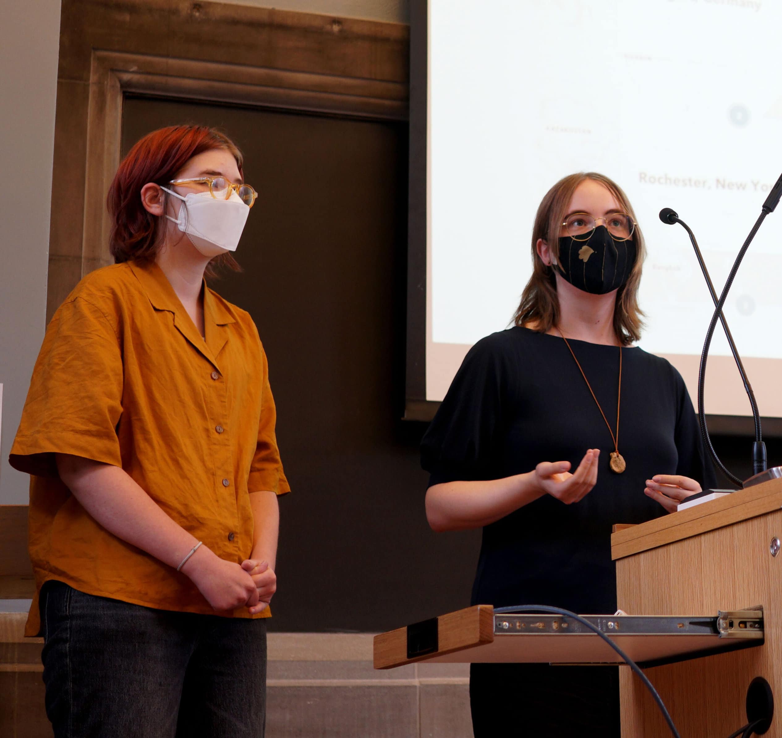 Izzy Friesen and Mies Kerr, wearing face masks, stand at a podium and give a presentation.