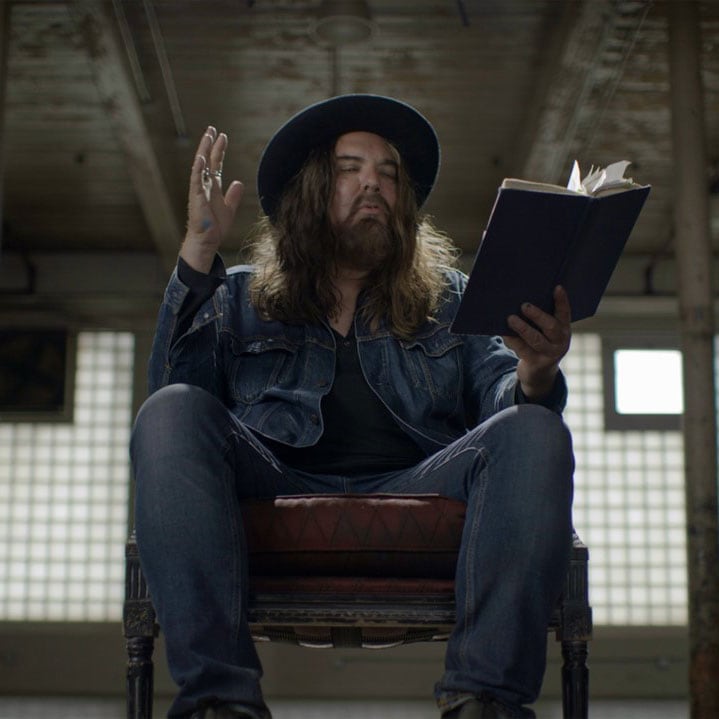 In a still from Beautiful Scars, Tom Wilson gestures as he sits in an old chair, holding a book.