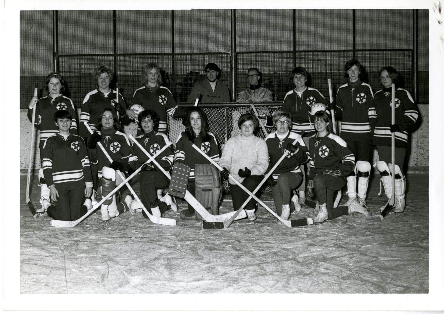 In an old photo from the 1960s, a women's hockey team poses in uniform on an ice rink.
