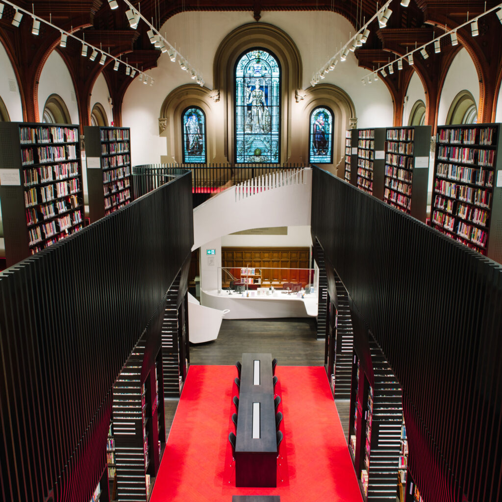 In the University College library, bookcases line a two-storey room with an arched stained glass window and a wooden ceiling.