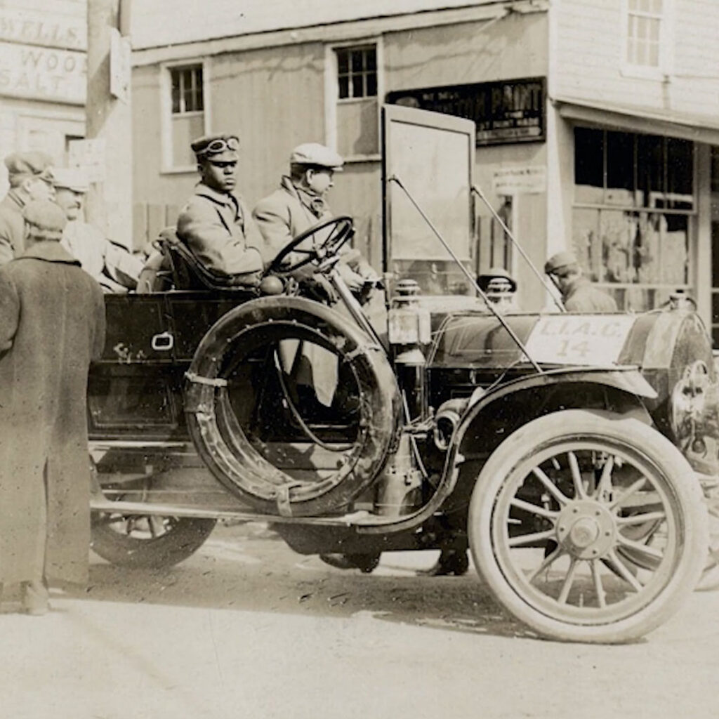 A Black man in chauffeur uniform sits at the wheel of an early automobile.