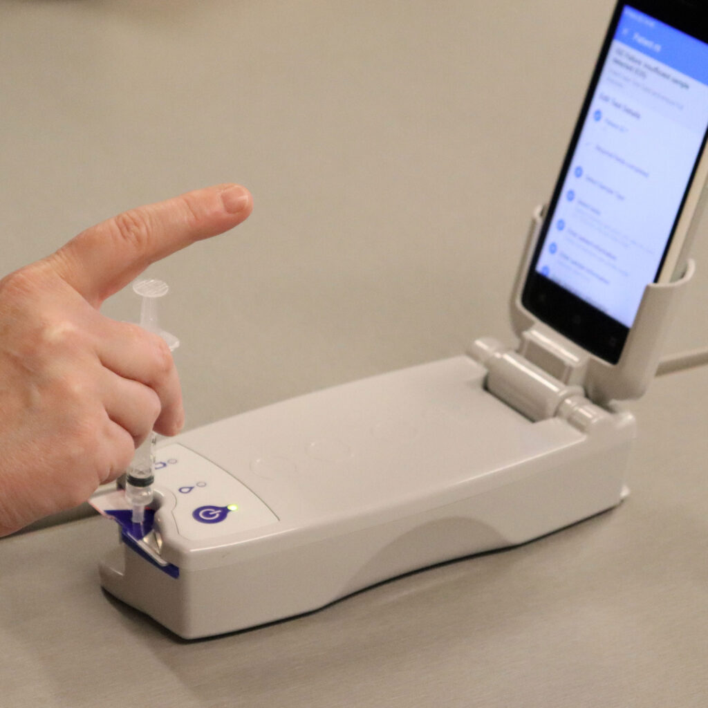 A hand inserts a syringe in a portable blood gas analysis system: a hand-sized device with a phone attached.