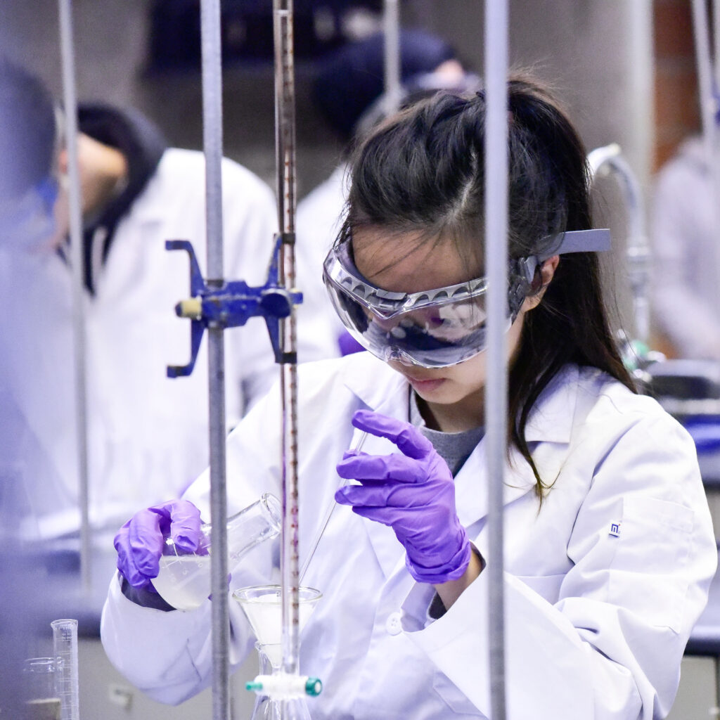 A young Asian woman measures a liquid in a chemistry class. She is wearing lab coat, gloves and safety glasses.