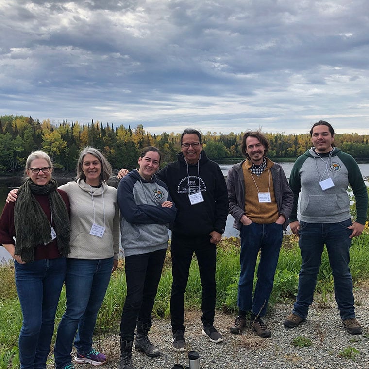 Indigenous knowledge holders and U of T professors smile, standing on the banks of the beautiful Rainy River.