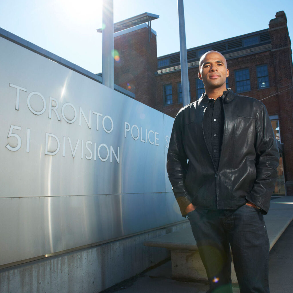 Akwasi Owusu-Bempah stands by a sign. Text reads: Toronto Police 51 Division.