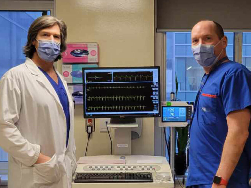 Michael Gollob and Mike Walker, wearing masks, stand by a heart monitoring console.