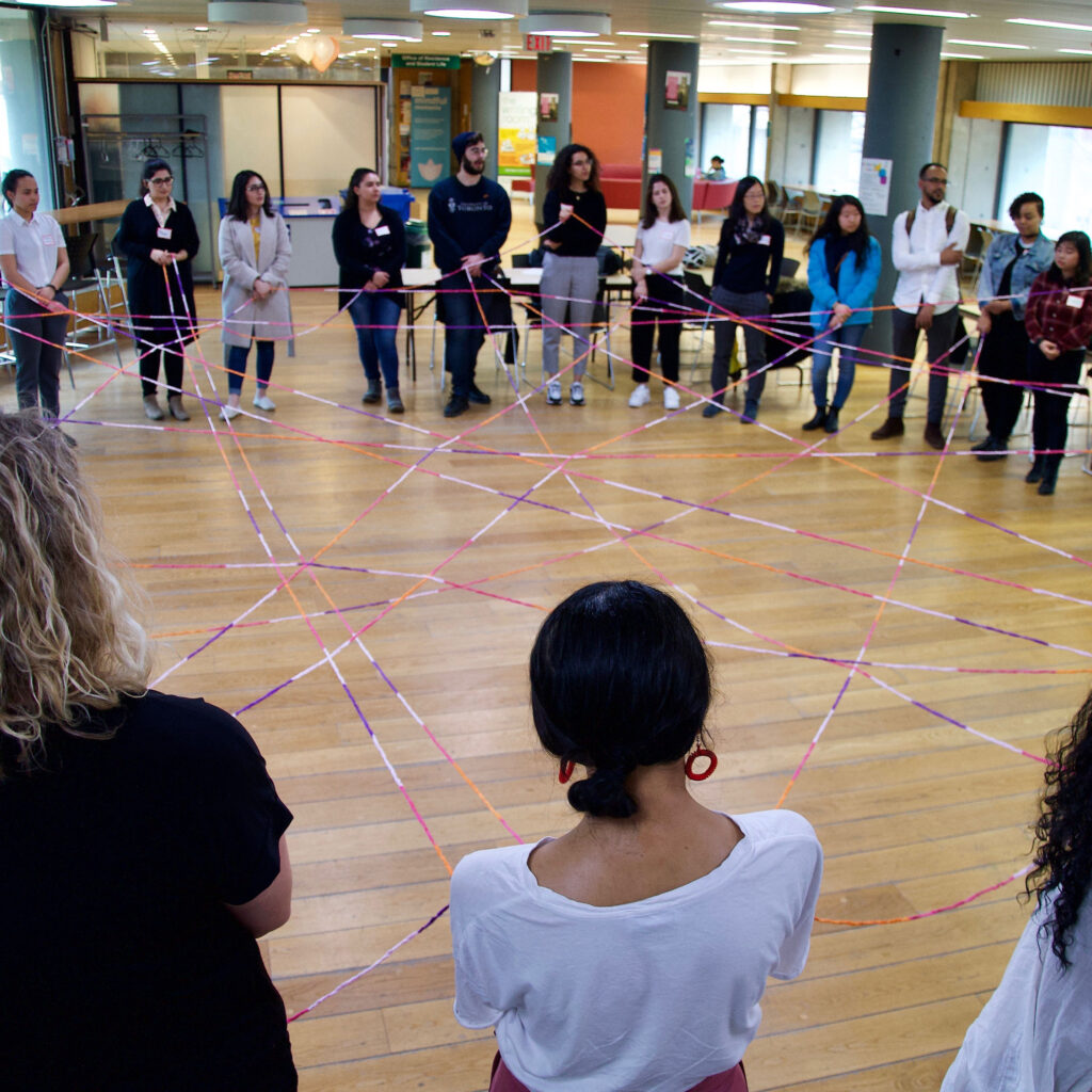 A group of people stand in a circle and hold colourful ropes that criss-cross the circle and connect them.