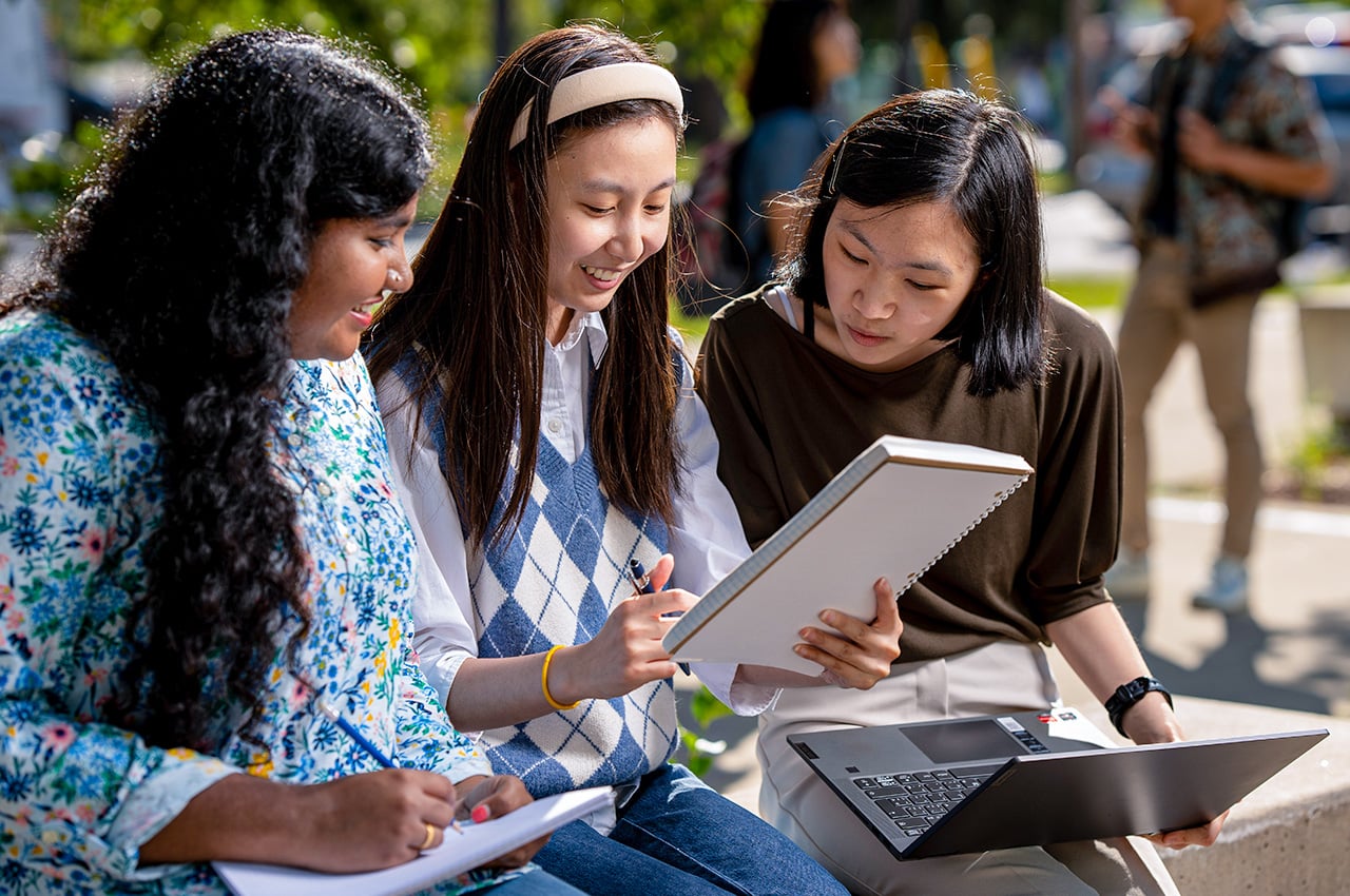 Three students compare notes while sitting on a wall outside.
