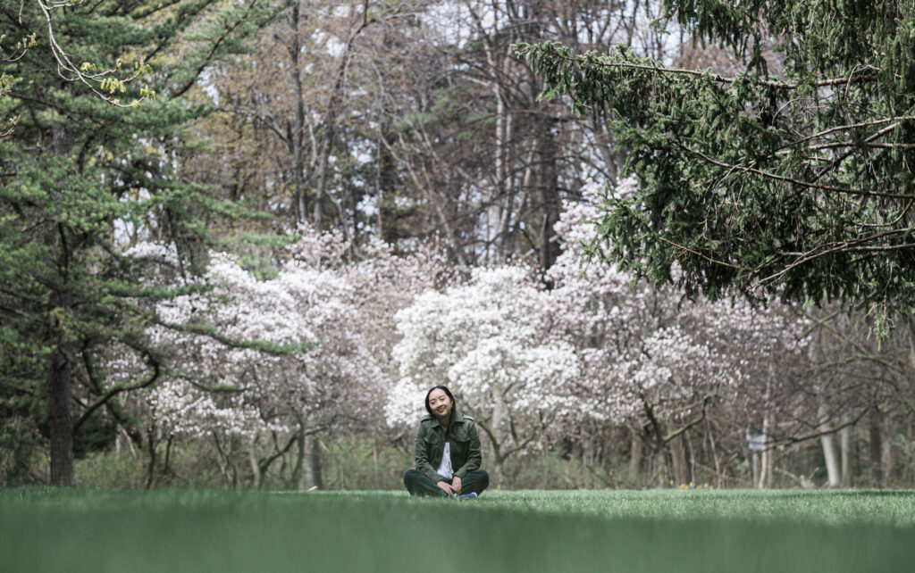 Amélie Desroches smiling and sitting on a green lawn, surrounded by blossoming trees.