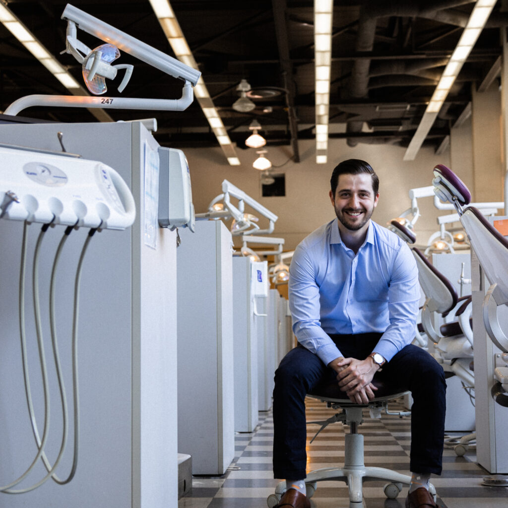 Dr. Michael Silva smiling. He is in the dental training clinic, with a row of dental chairs in cubicles.
