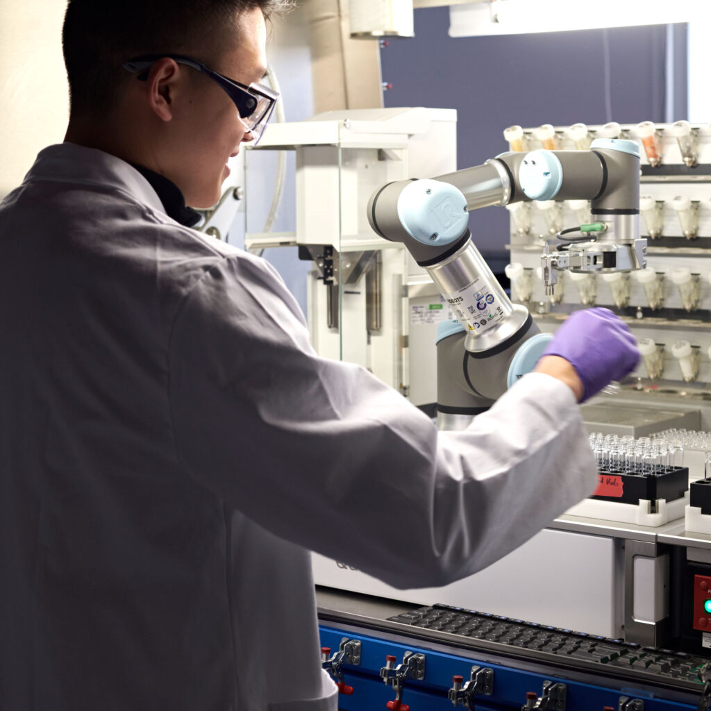 A man in lab coat, gloves and goggles stands at a lab bench and lifts a small glass vial from a machine