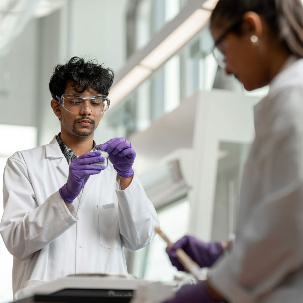 A man in goggles, gloves and lab coat opens a small jar in a sunny laboratory.