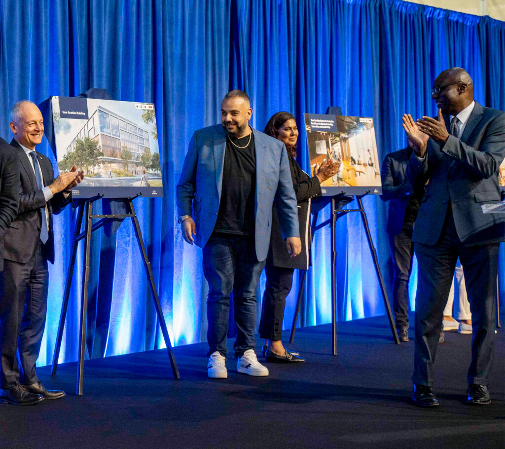 A group of men and women stand on a stage, clapping, with renderings of new buildings behind them