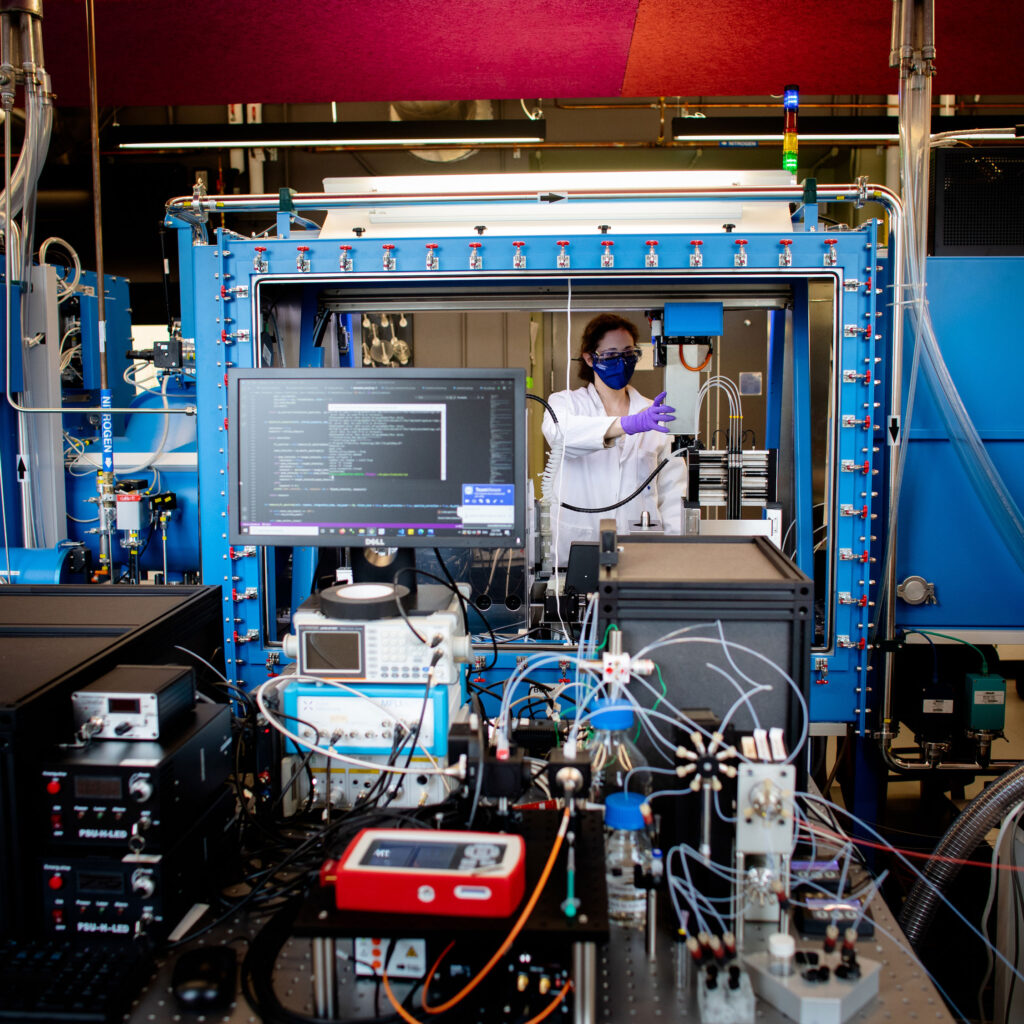 Emily Hopkins, in mask and gloves, operates a materials acceleration platform, a large steel case wired to many computers.