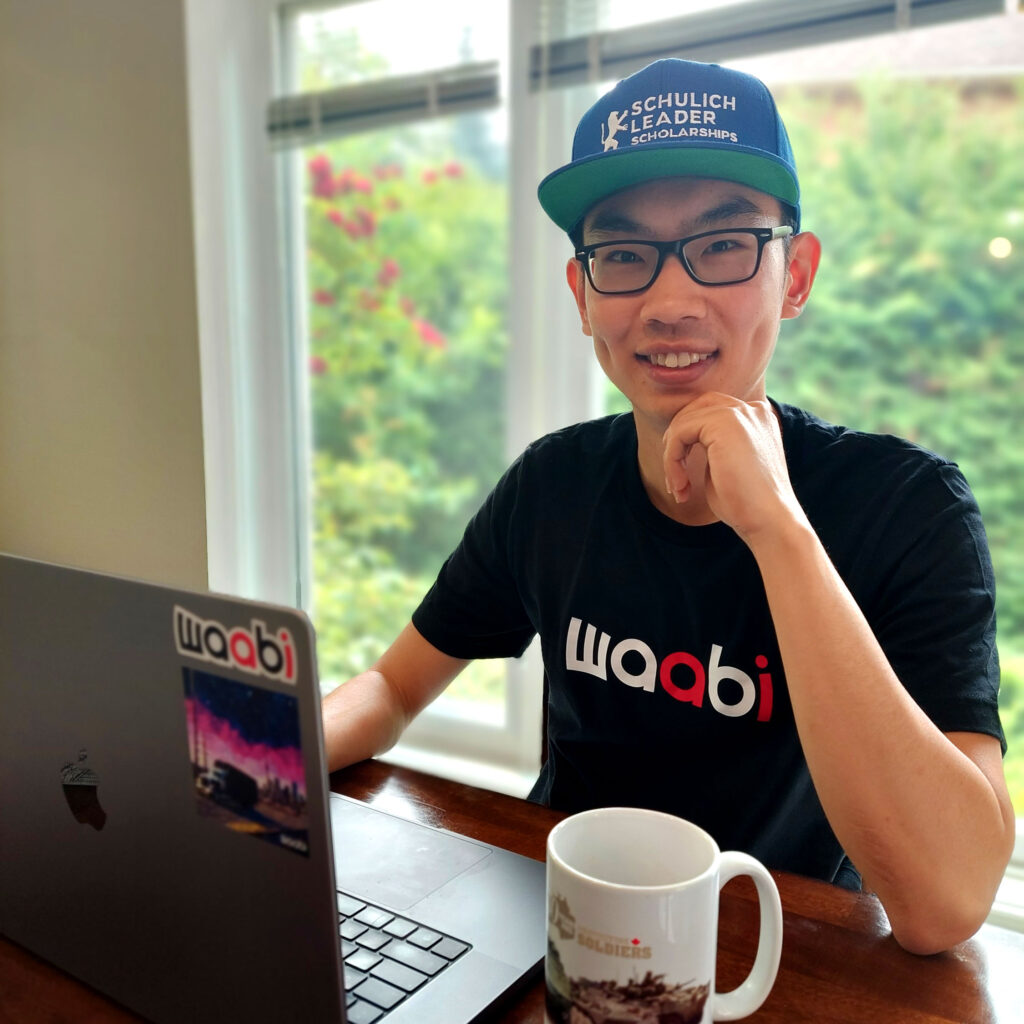 James Xu working at a laptop in front of a window. He wears a Waabi t-shirt and a Schulich Leader baseball cap.