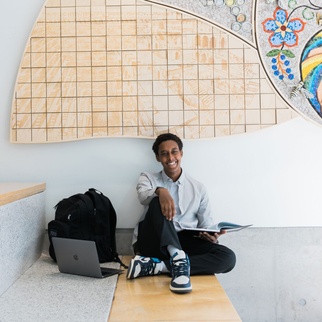 Badri Widaatalla smiling and sitting with backpack and laptop under a colourful mural of the fibonacci sequence.