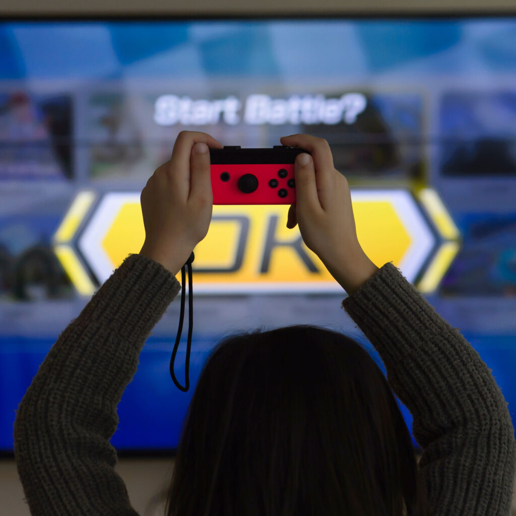 A young woman holds a video game controller over her head as she faces a screen with the text: Start Battle?