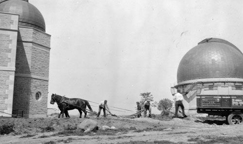 In an image from 1934, two horses pull ground-levelling equipment. A domed observatory roof stands behind them.