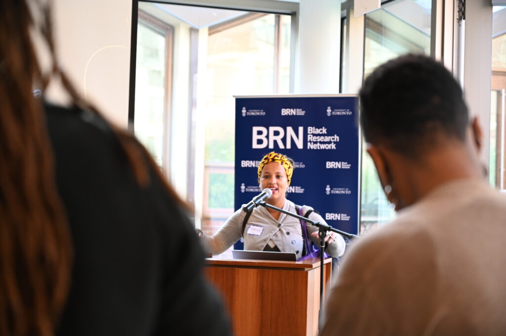 A young Black female research stands at the podium speaking at a BRN Welcome Social event.